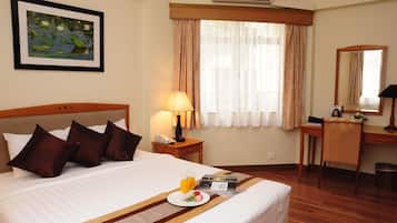 A hotel room with a neatly made bed, a bedside table with a lamp, a desk with a chair, a large window with curtains, and a framed picture on the wall.
