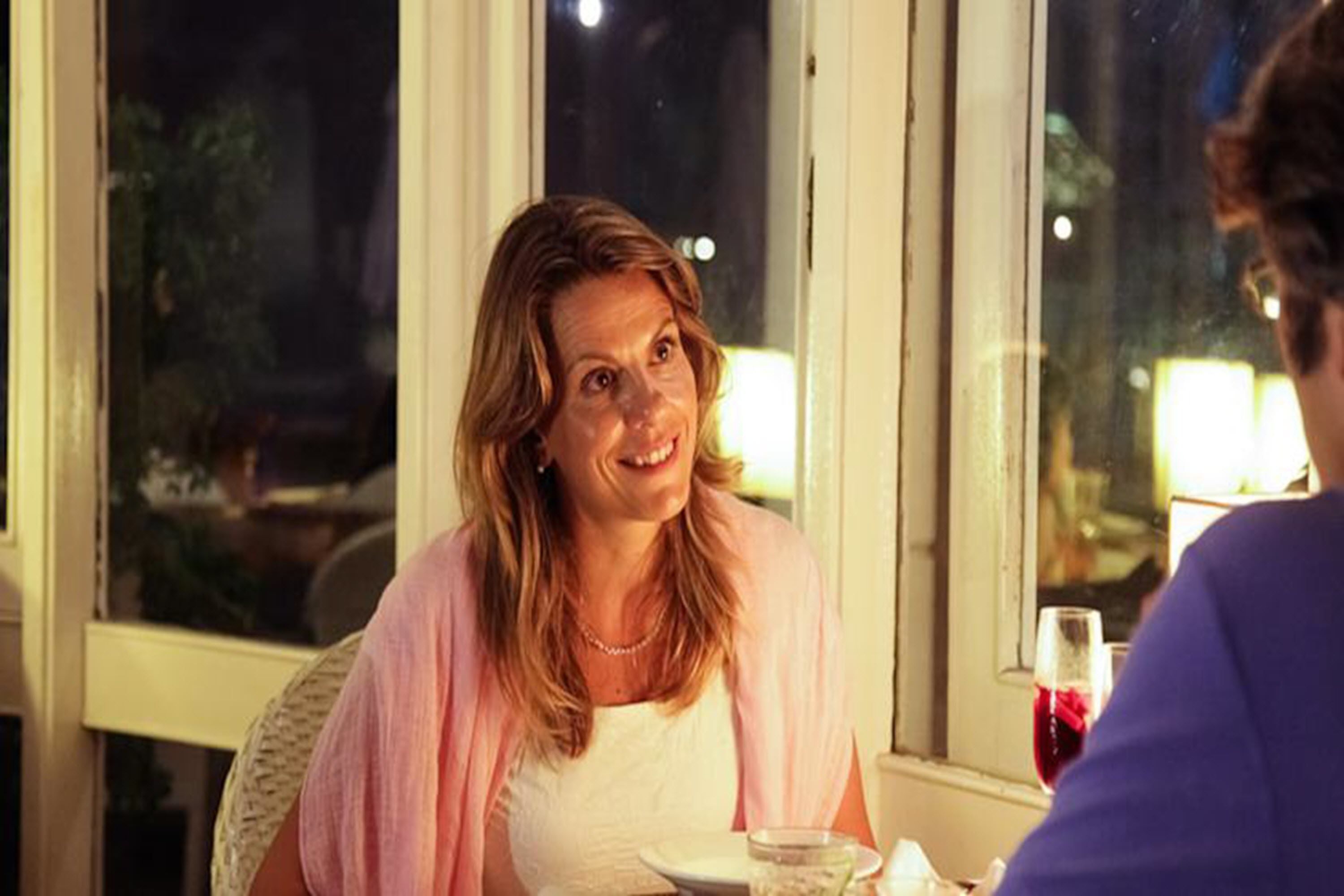 Une personne est assise à une table, devant un verre de vin rouge, dans une pièce où l’on aperçoit une fenêtre et une porte.