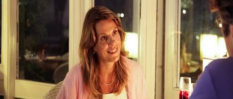 Une personne est assise à une table, devant un verre de vin rouge, dans une pièce où l’on aperçoit une fenêtre et une porte.