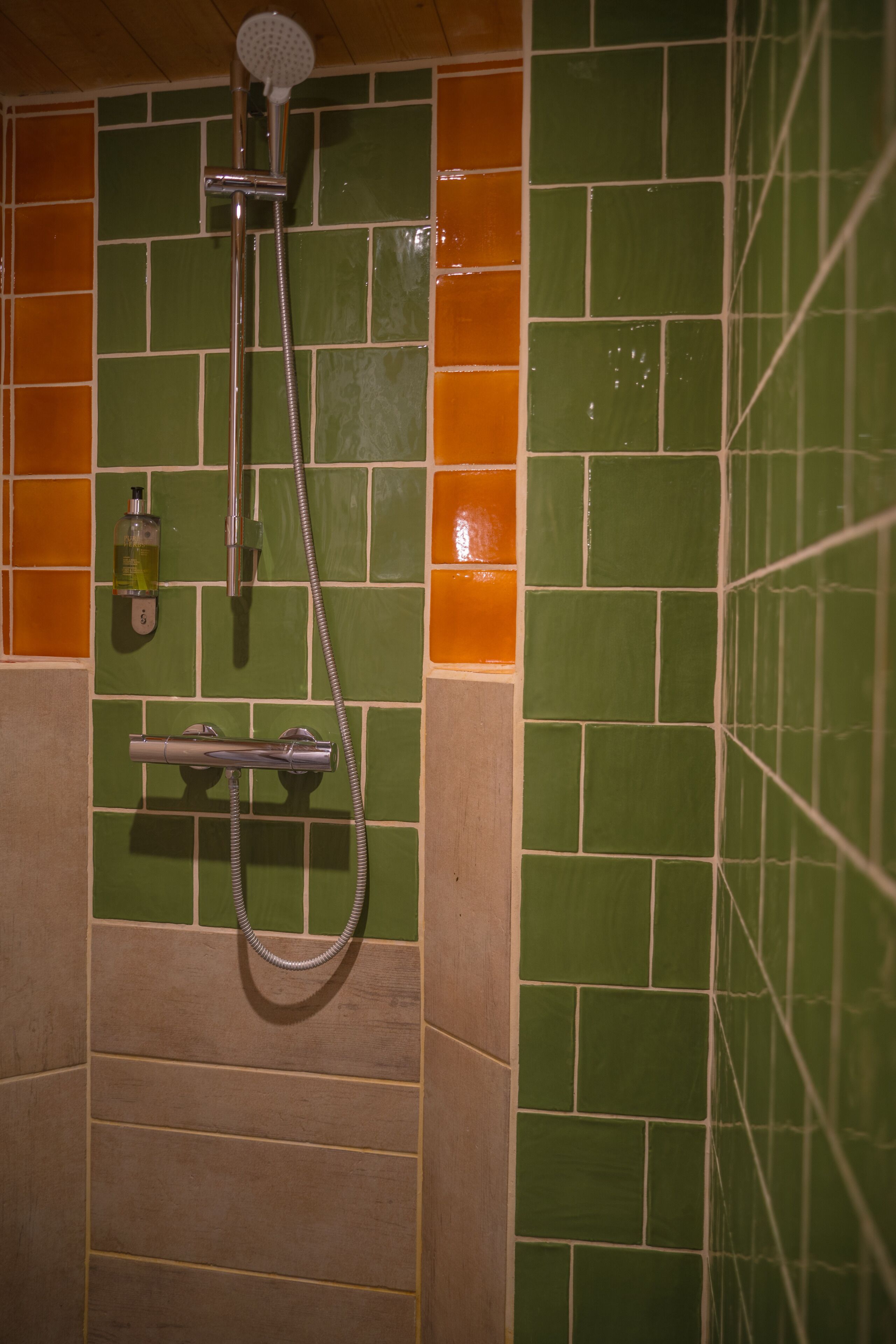 Un área de ducha con azulejos verdes y anaranjados, un cabezal de ducha de mano y un dispensador de jabón.