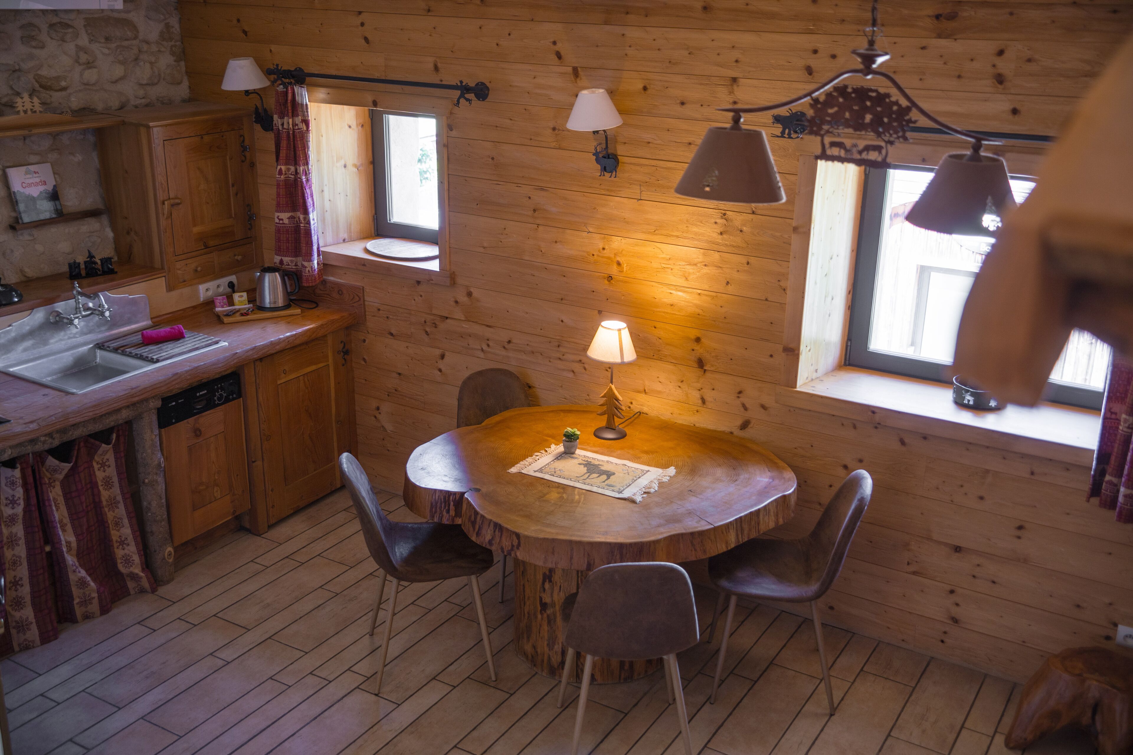Een houten blokhutinterieur met een ronde eettafel, stoelen, een keuken met een spoelbak en kastjes, en twee ramen met gordijnen.