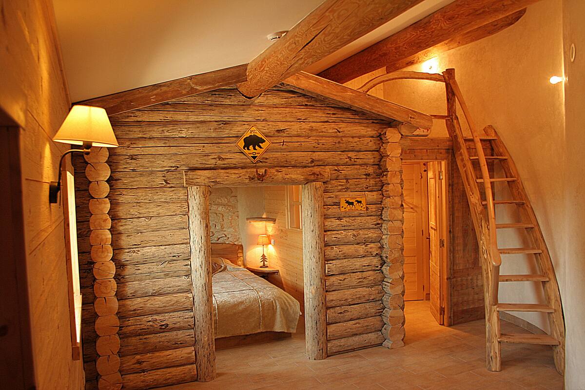 A log cabin-style room with a bed, a nightstand with a lamp, a wooden ladder, and a bear warning sign.