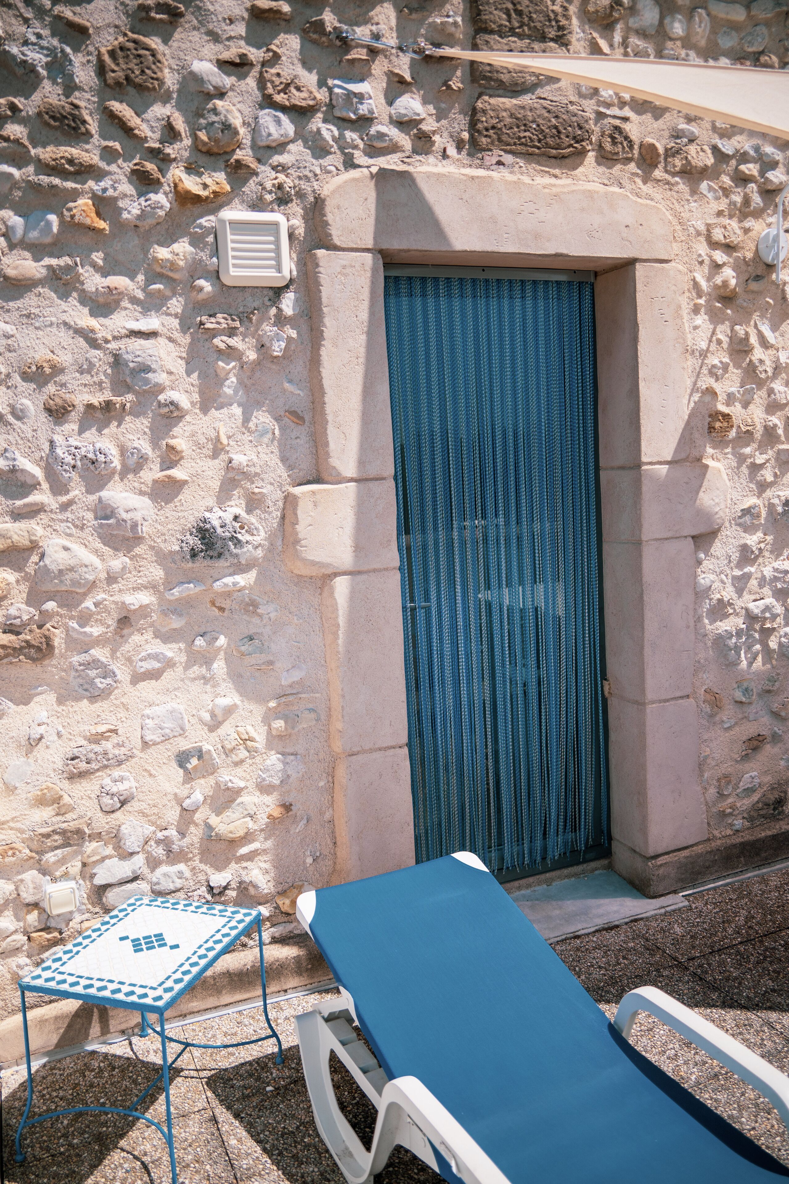 Un muro de piedra con una puerta azul, una mesa pequeña y un sillón azul.