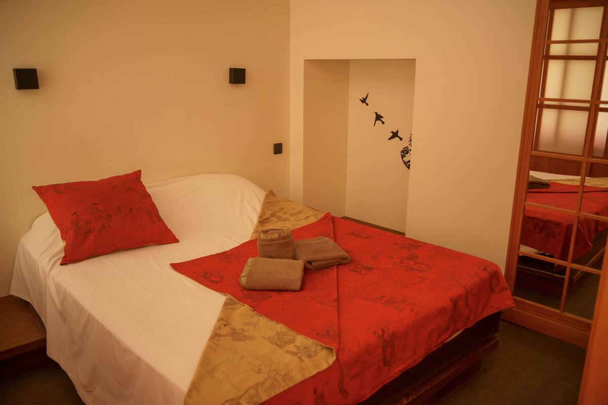 A neatly made bed with red and beige bedding, a red pillow, and a beige throw blanket.