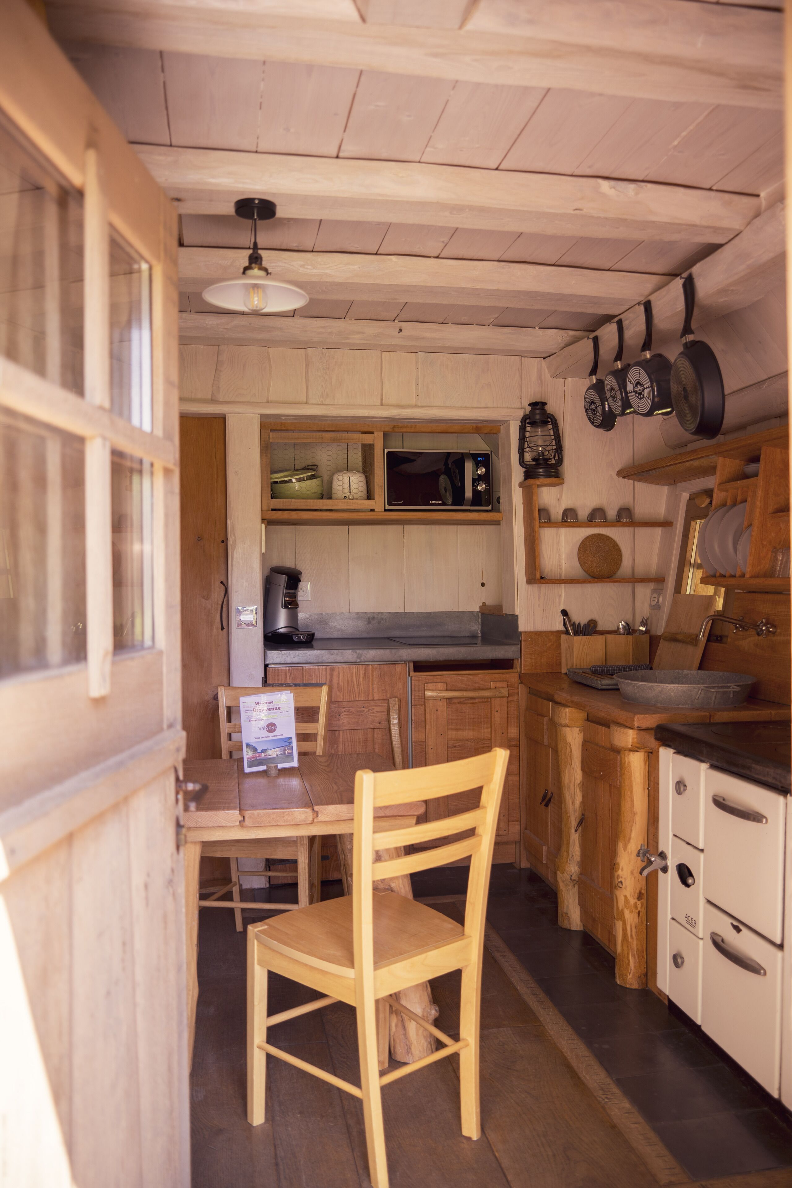 Un intérieur en bois comprenant une table et des chaises, un coin cuisine avec des placards et une cuisinière, ainsi qu’un luminaire suspendu.