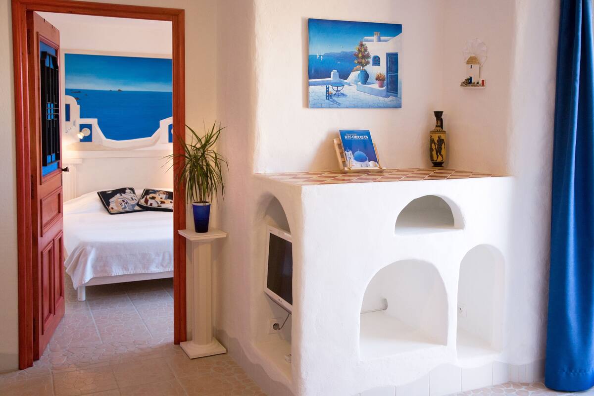 A room with a bed, a small table with a book, a potted plant, and a painting of a coastal scene.