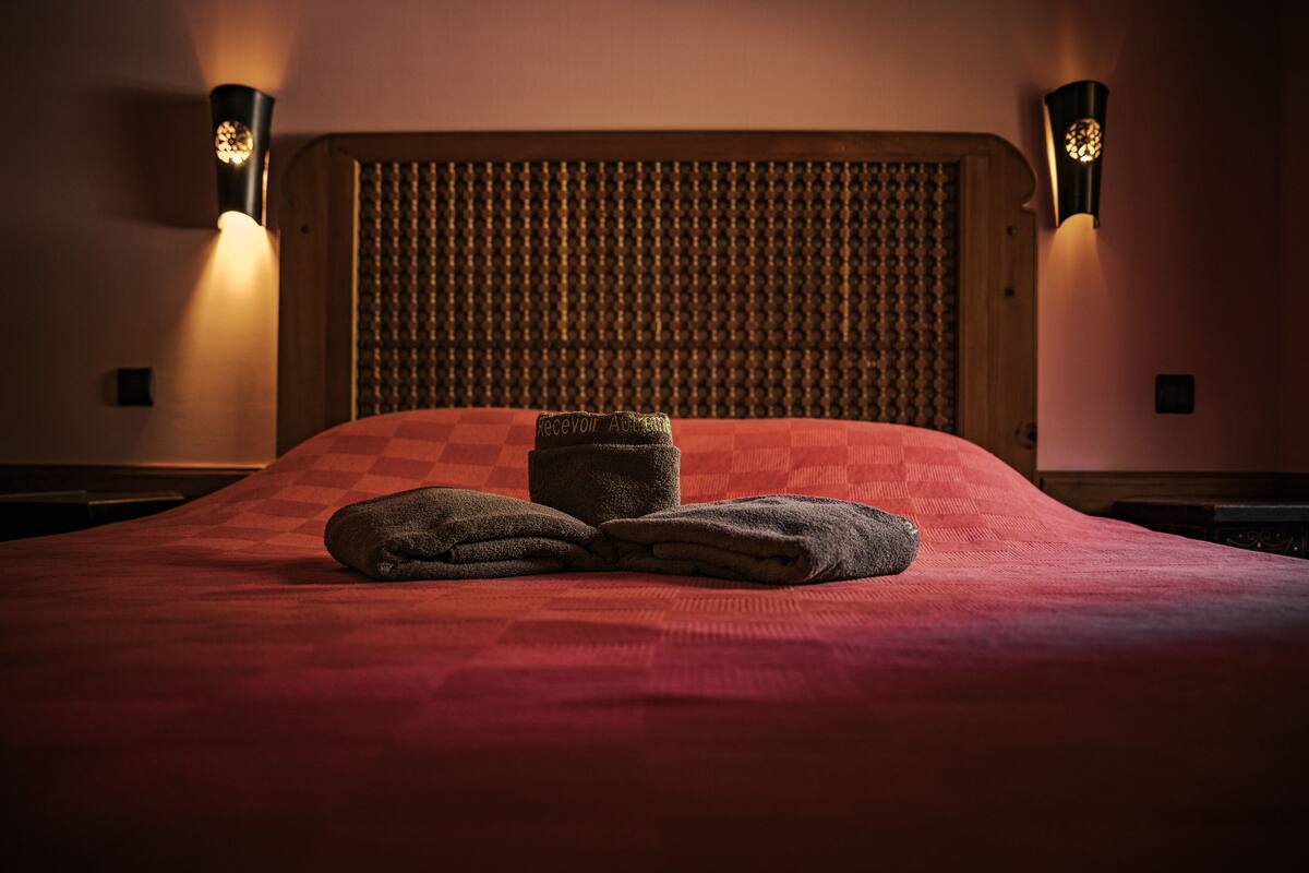 A hotel room with a checkered red bedspread, a wooden headboard with a textured panel, and two wall-mounted lamps on either side.