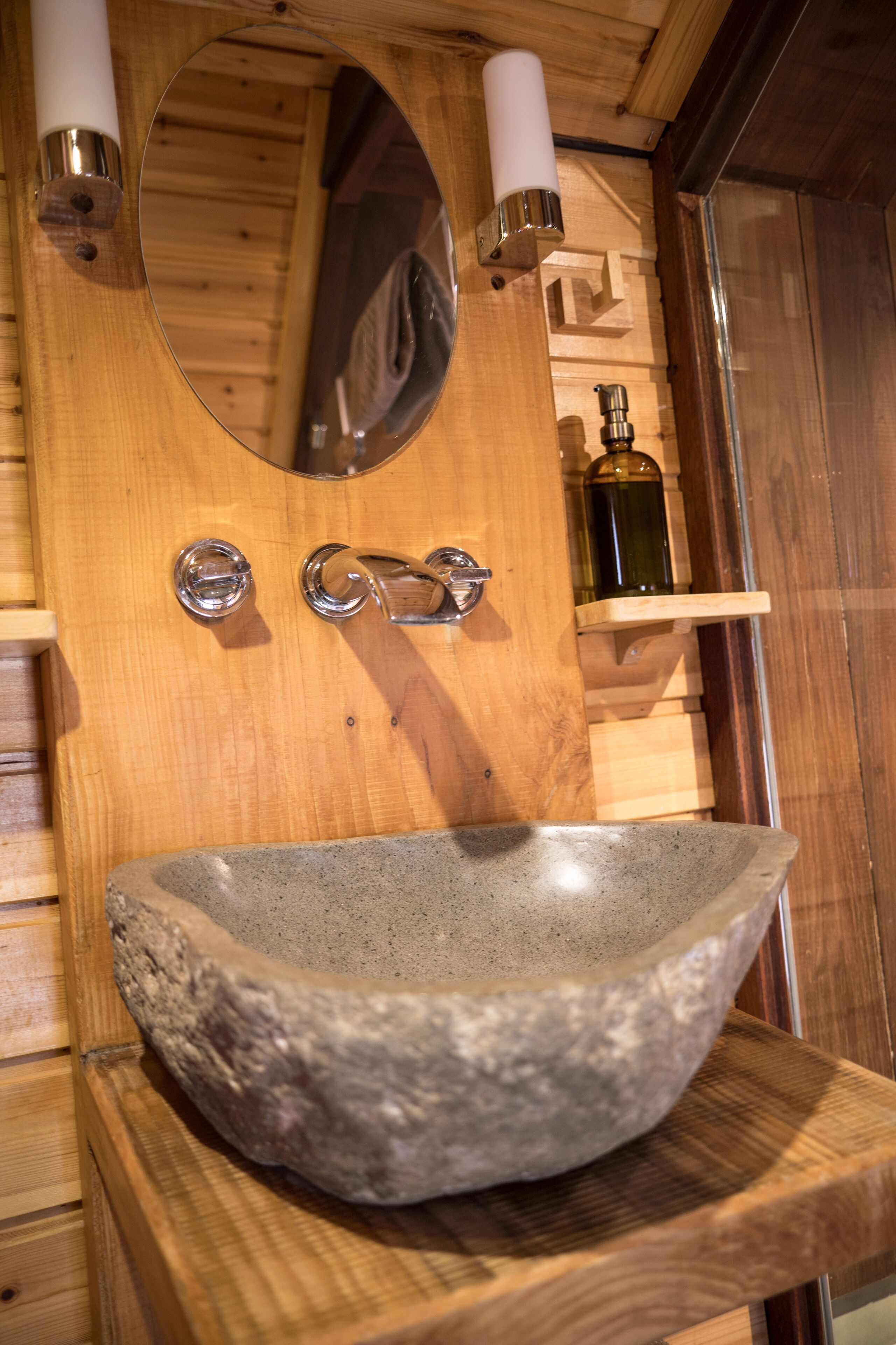 A stone basin sink with a wooden countertop, a wooden wall with a mirror, and a shelf with a bottle.