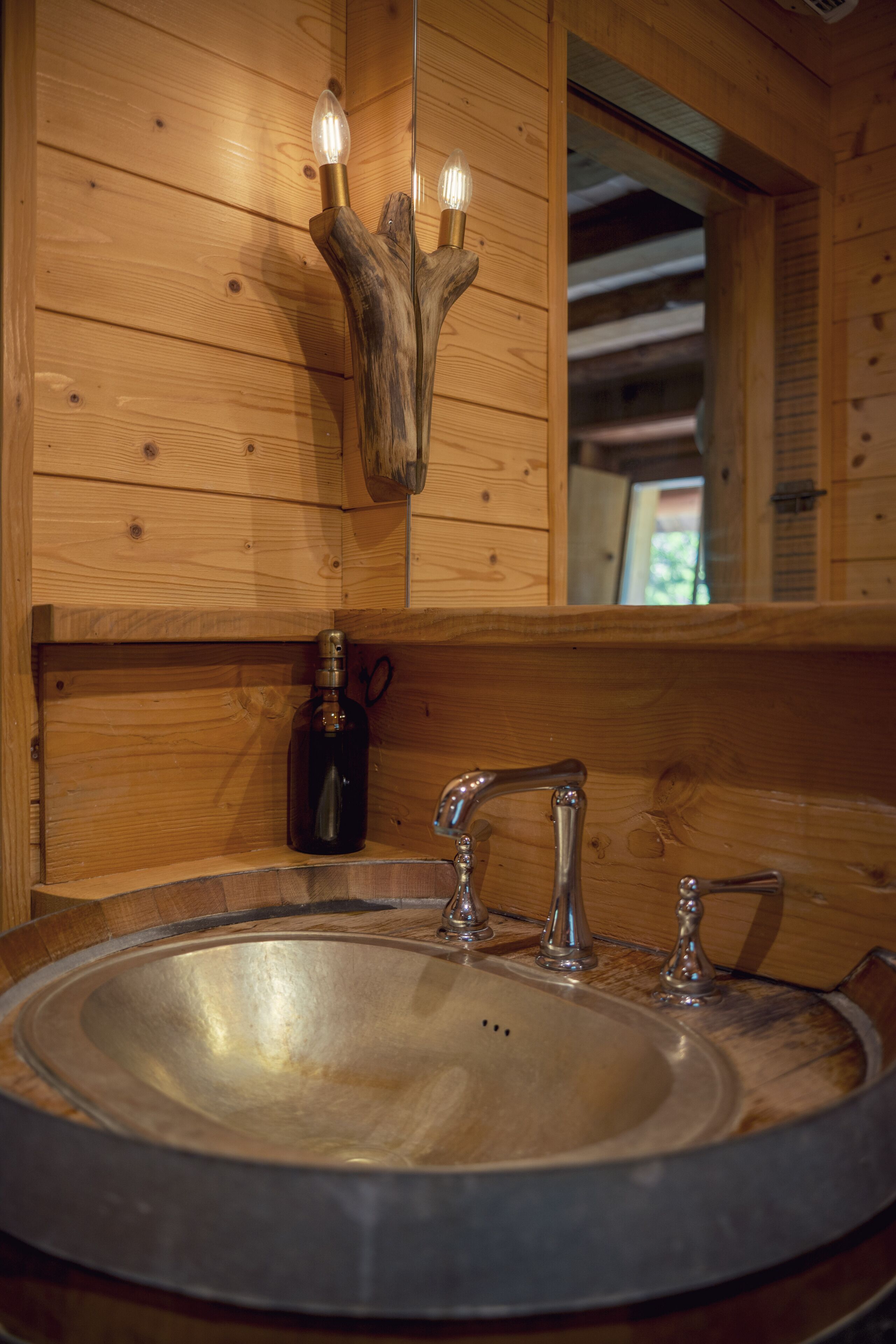 A wooden cabin interior with a round sink, a wooden wall-mounted light fixture, and a mirror reflecting the cabin's interior.
