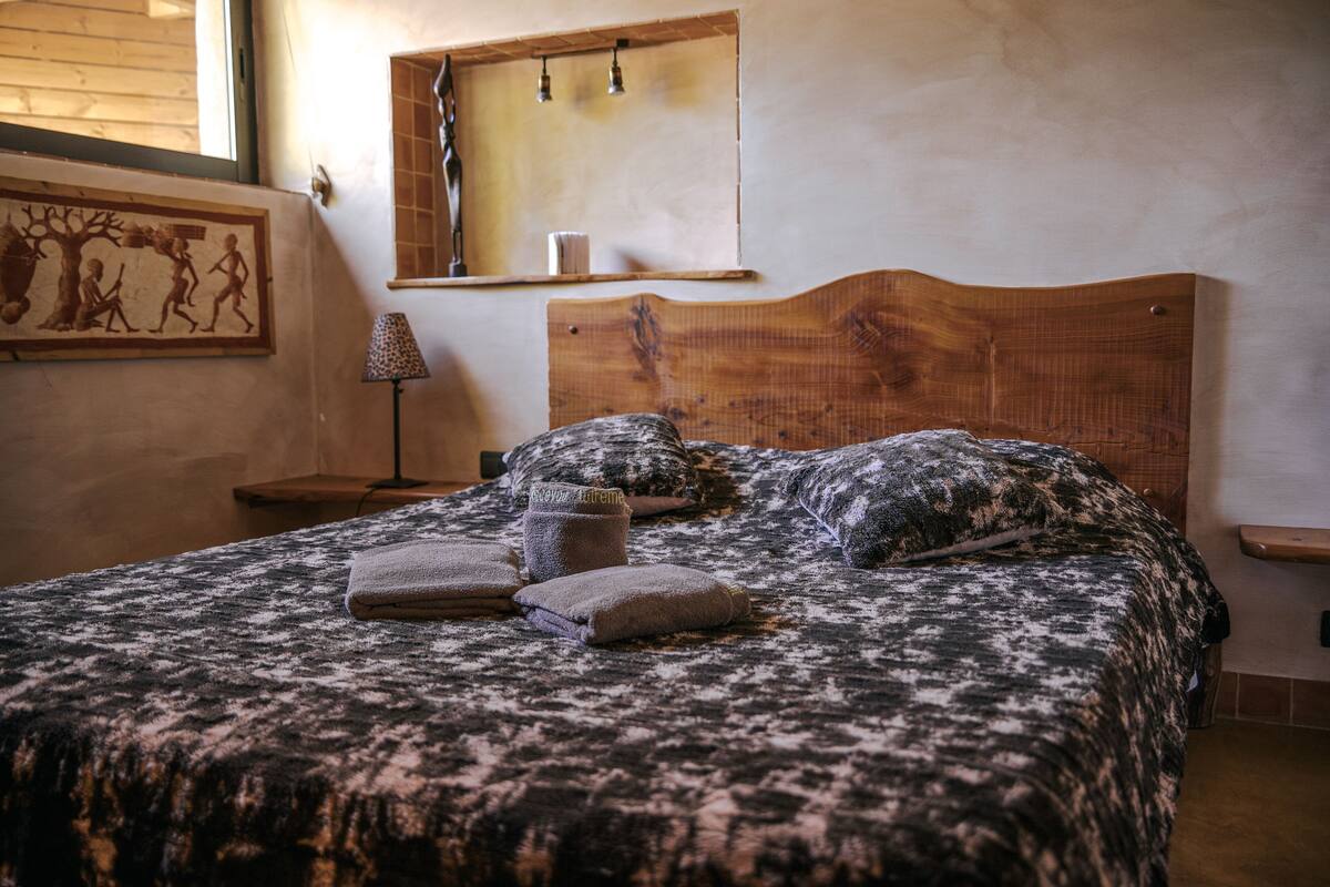A neatly made bed with a patterned bedspread, a wooden headboard, and a wall-mounted shelf with a mirror and a small lamp.