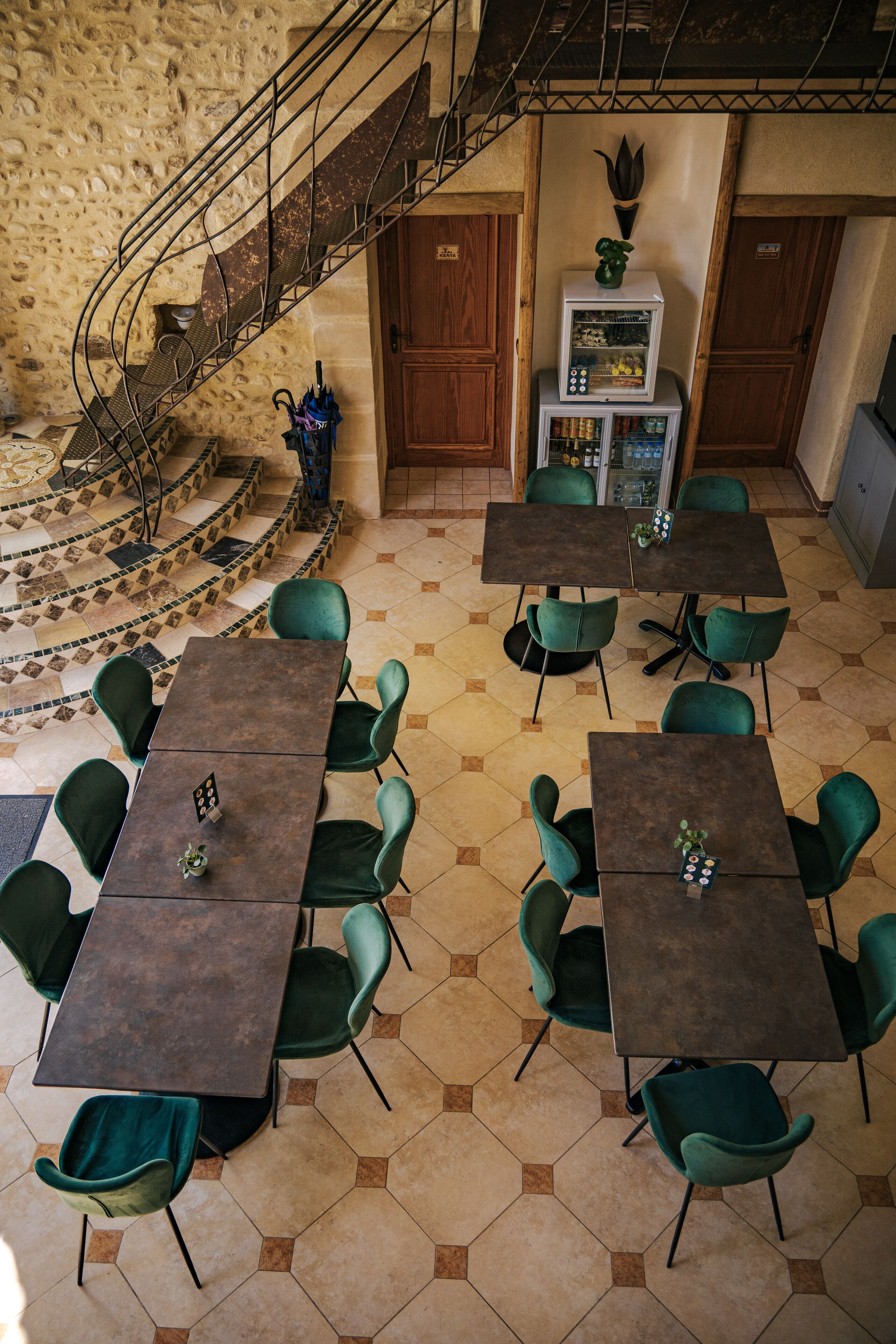 A restaurant with hexagonal tiled flooring, green upholstered chairs, and rectangular tables. A spiral staircase is visible in the background.