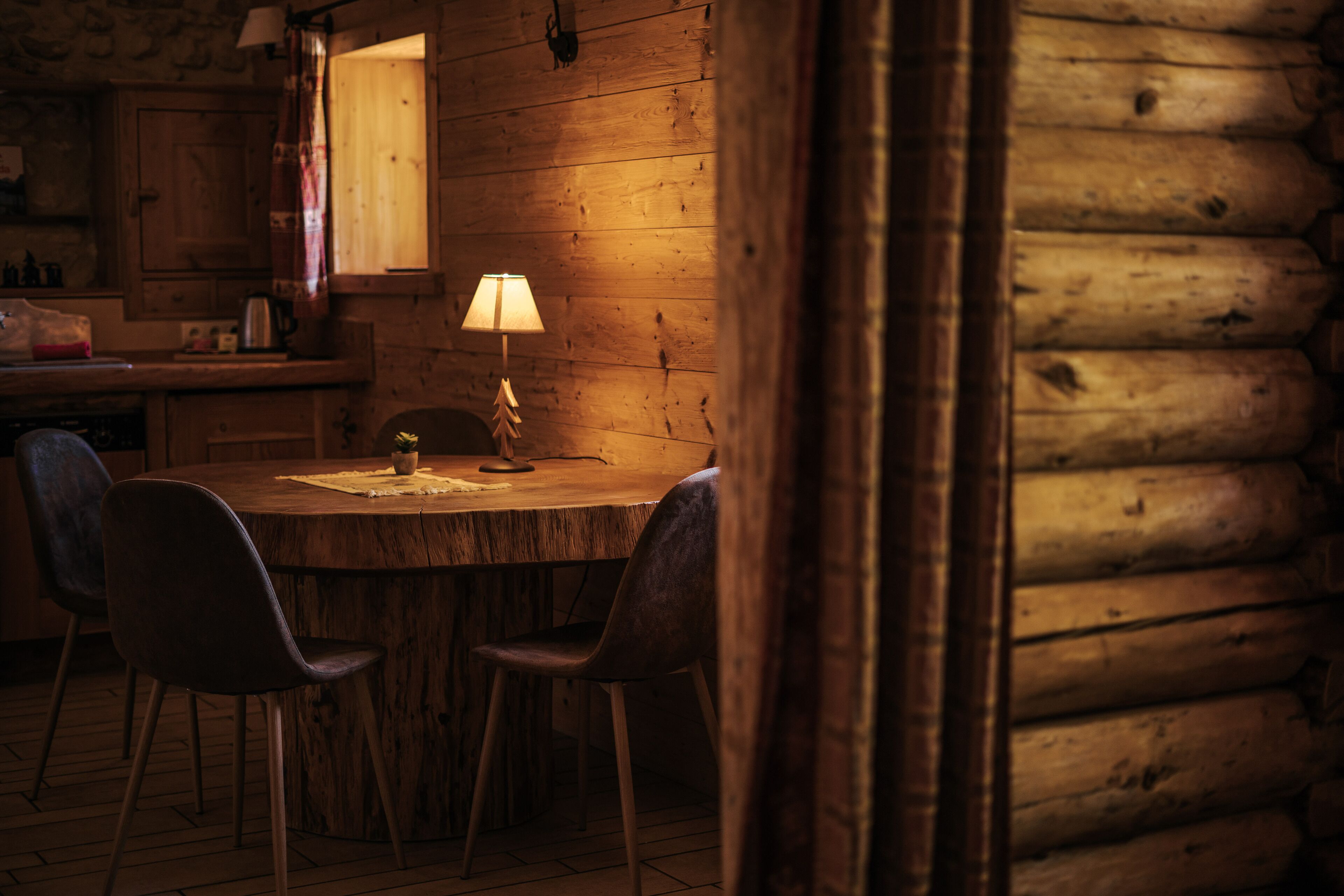 Un intérieur rustique en bois, avec une table de salle à manger ronde, deux chaises, une lampe et une fenêtre avec des rideaux.