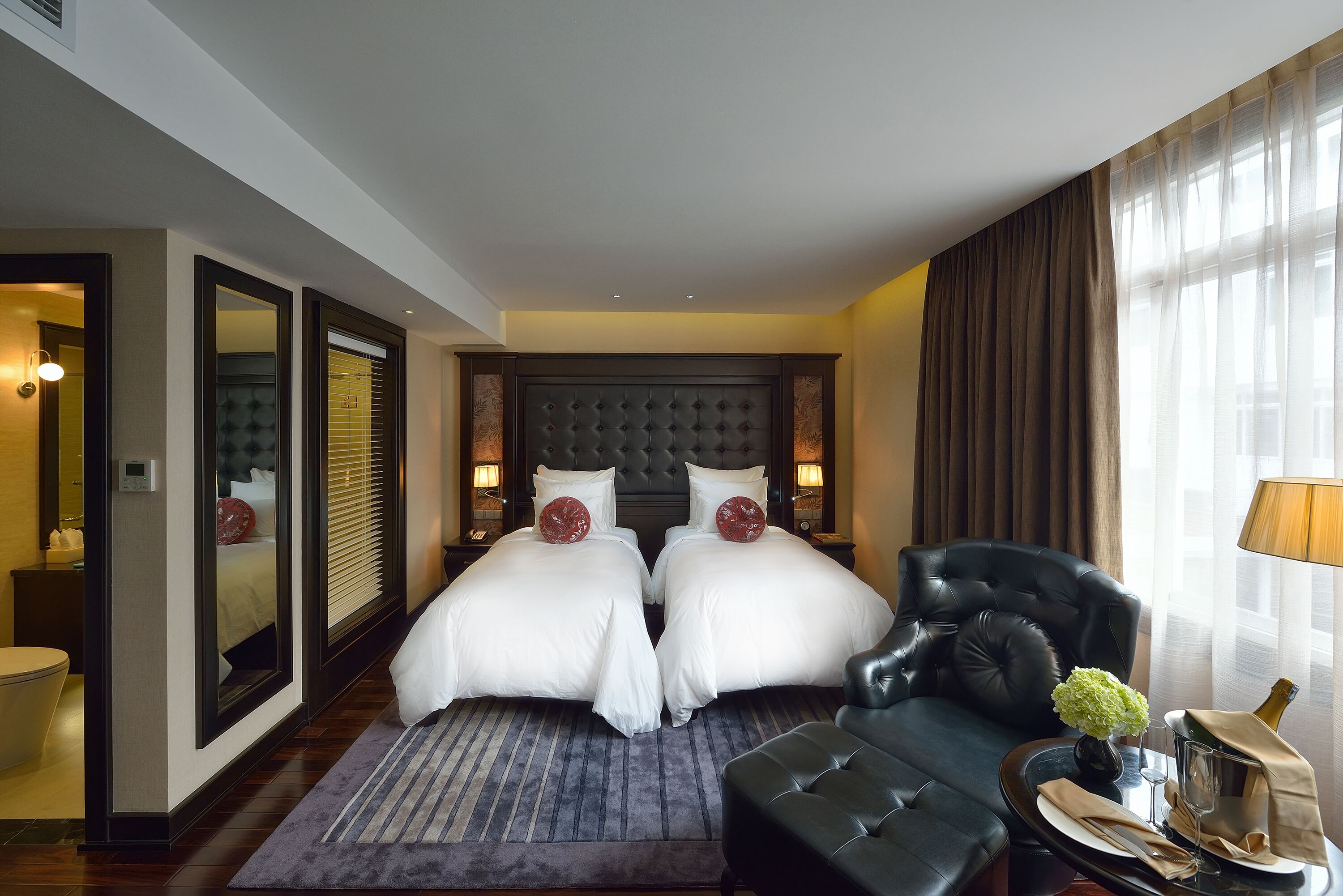 A hotel room with two beds, a sofa, a small table with a vase and champagne, and a view of the city through large windows.