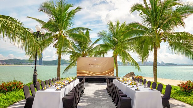 Een buitenterras met een lange tafel en stoelen gericht op een schilderachtig uitzicht over de oceaan, met palmbomen en een groot scherm waarop evenementinformatie staat.