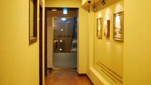 Un couloir avec une porte portant l’inscription « CUISINE » qui donne sur une pièce. Des tableaux encadrés ornent le mur, et une pancarte avec du texte y est également fixée.