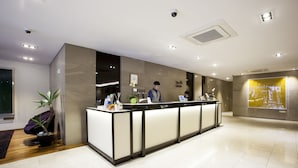 A hotel reception area with a reception desk, two staff members, a potted plant, and a large digital screen displaying a cityscape.