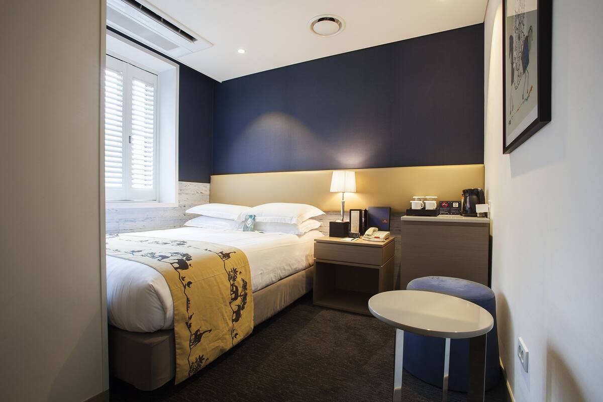 A hotel room with a bed, bedside table, lamp, and a small round table.