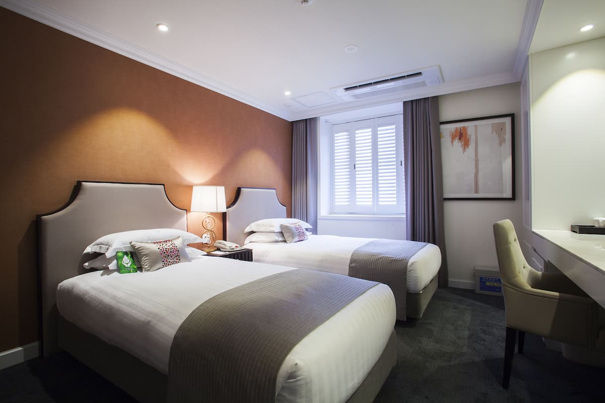 A hotel room with two beds, a desk, and a chair. There is a window with curtains, a lamp, and a framed picture on the wall.