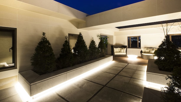 A modern outdoor area with a tiled floor, a row of evergreen trees, and a seating area with a sofa and chairs.