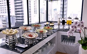 A buffet setup with various food items and drinks on a counter by a large window overlooking a cityscape. - Carina Hotel (Nha Trang)