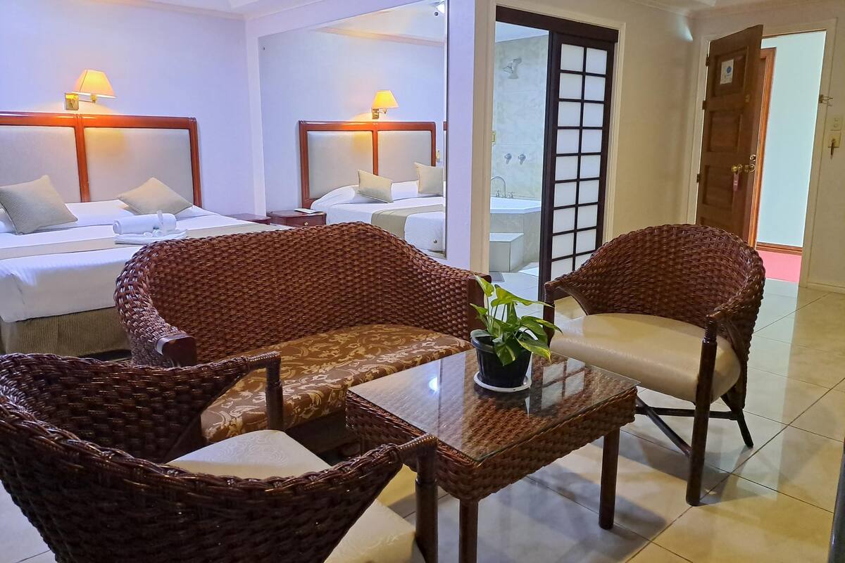 A hotel room with two beds, wicker chairs, a glass table, and a potted plant.