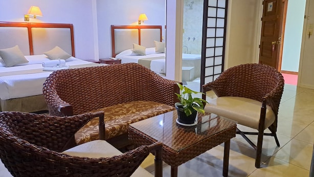 Habitación de hotel con dos camas, sillas de mimbre, una mesa de vidrio y una planta en maceta.