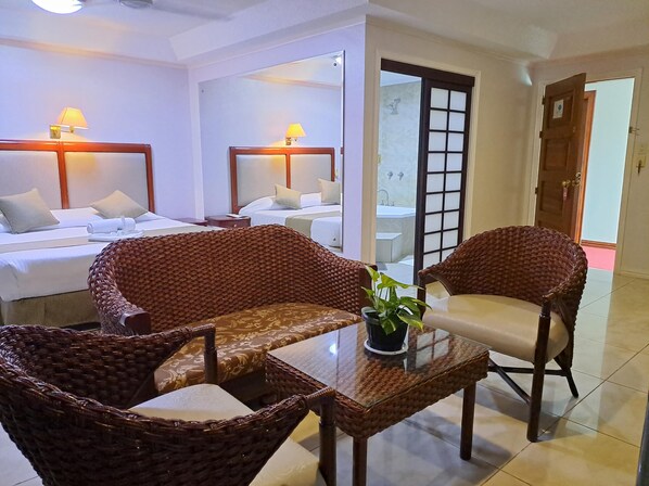 Habitación de hotel con dos camas, sillas de mimbre, una mesa de vidrio y una planta en maceta.