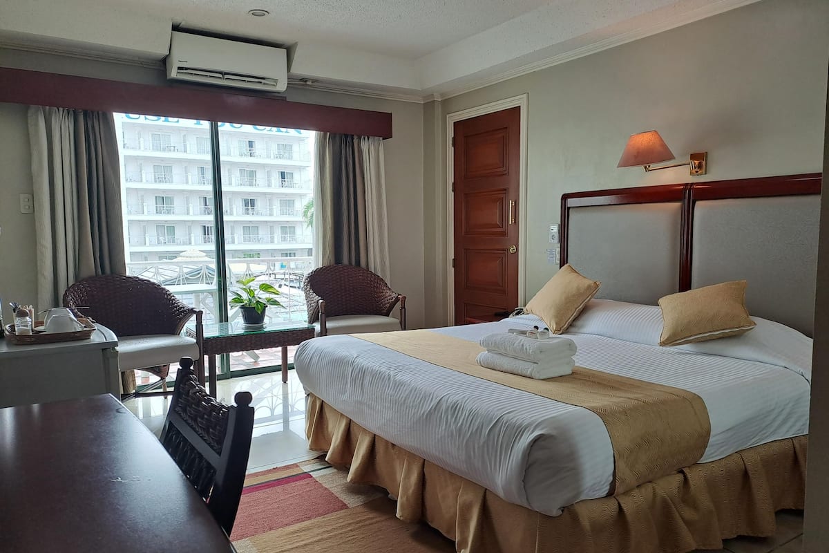 A hotel room with a bed, two chairs, a desk, and a view of a building through the window.