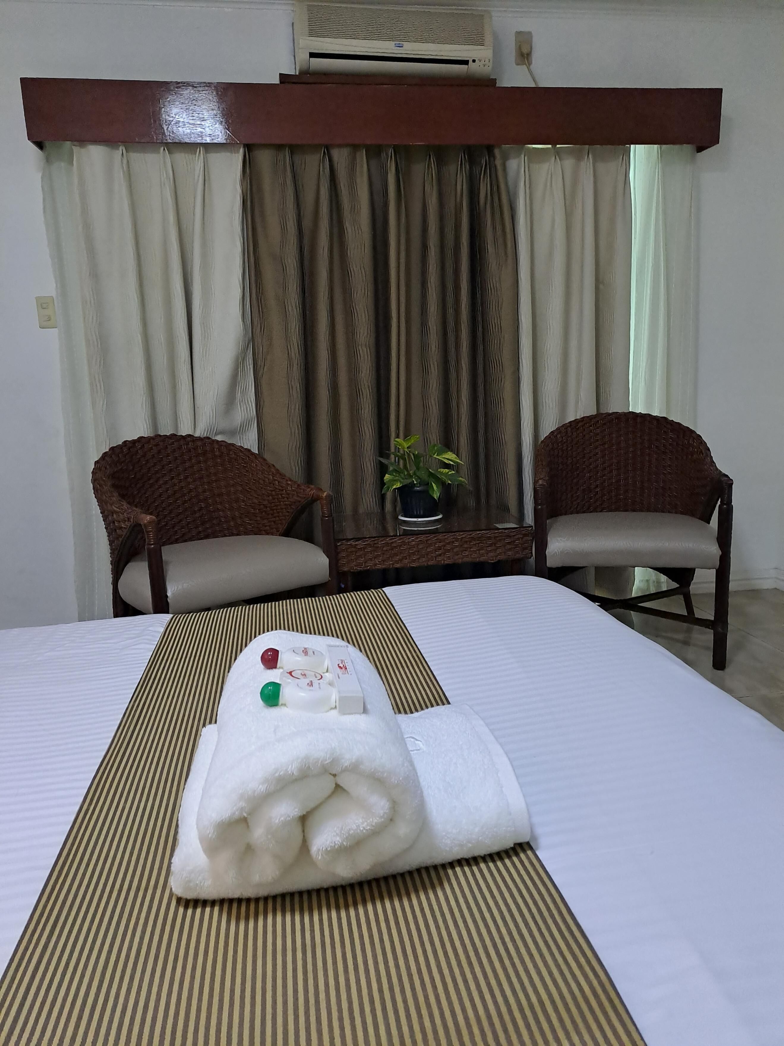 A hotel room with a bed, two wicker chairs, a small table with a potted plant, and a white towel on the bed.