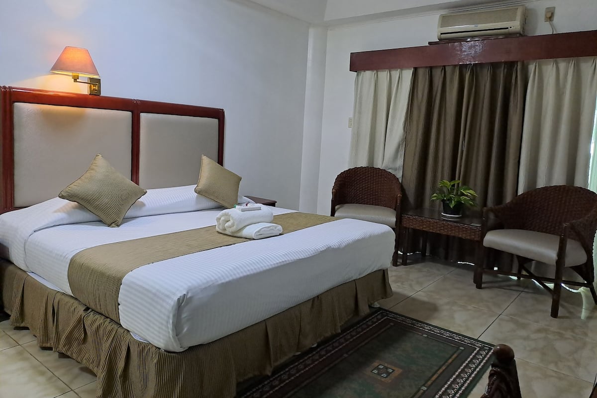 A hotel room with a bed, a nightstand, a lamp, a chair, a table, and an air conditioning unit.