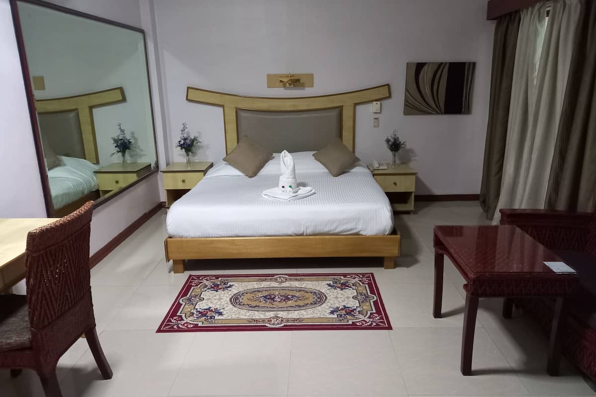 A hotel room with a large bed, a wooden headboard, a bedside table with a lamp, a chair, a desk, and a large mirror.