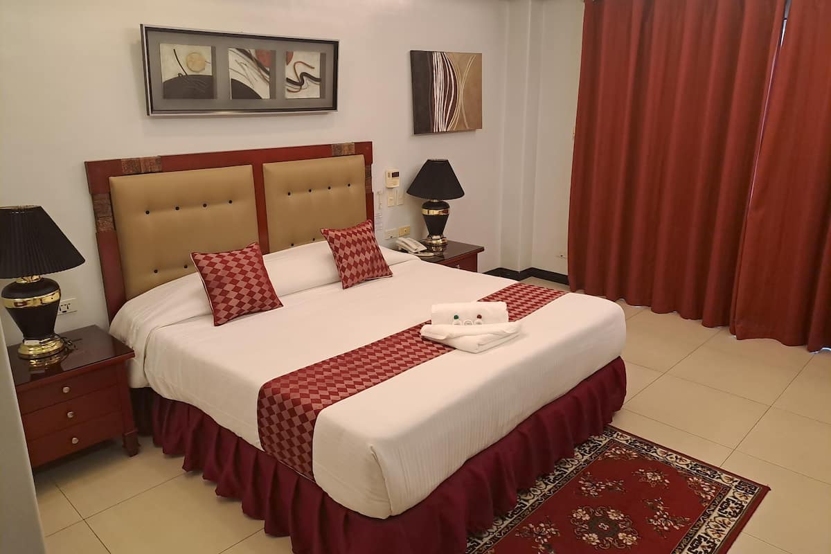 A neatly made bed with a checkered bedspread and pillows, a red curtain, and a framed abstract painting on the wall.