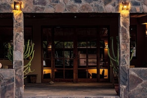 Un arco de piedra con puerta de vidrio, que da paso a una habitación con estanterías de madera y plantas de cactus.