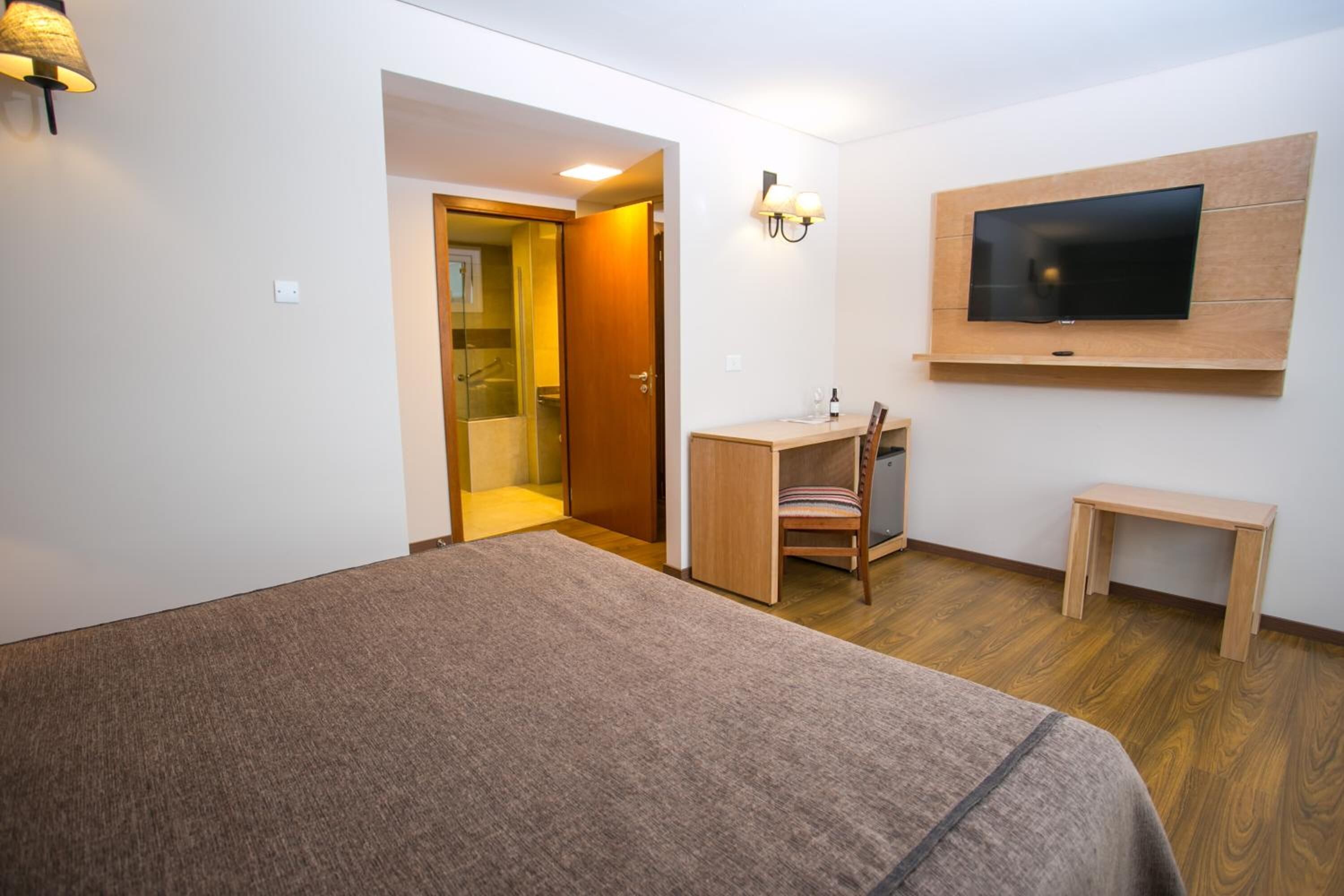 A bedroom with a bed, a desk, a chair, a TV mounted on the wall, and a door leading to a bathroom.
