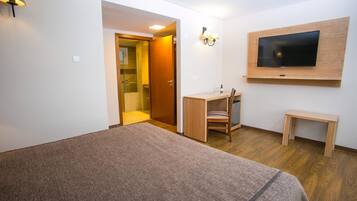 A bedroom with a bed, a desk, a chair, a TV mounted on the wall, and a door leading to a bathroom.
