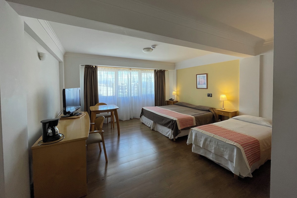 Ein Hotelzimmer mit Schreibtisch, zwei Betten, Fernseher und einem Fenster mit Vorhängen.