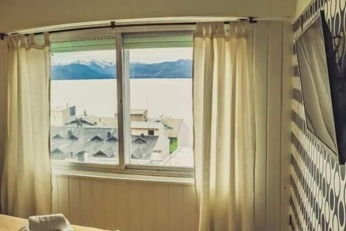 Habitación de hotel con una cama, dos lámparas de pared, una ventana con cortinas y vistas a edificios y montañas.