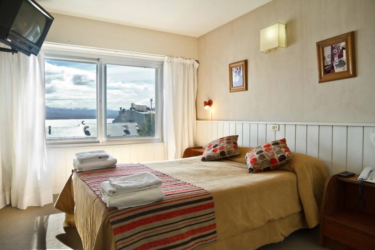 Habitación de hotel con cama, televisión, ventana con cortinas, mesita de noche y lámpara de pared.