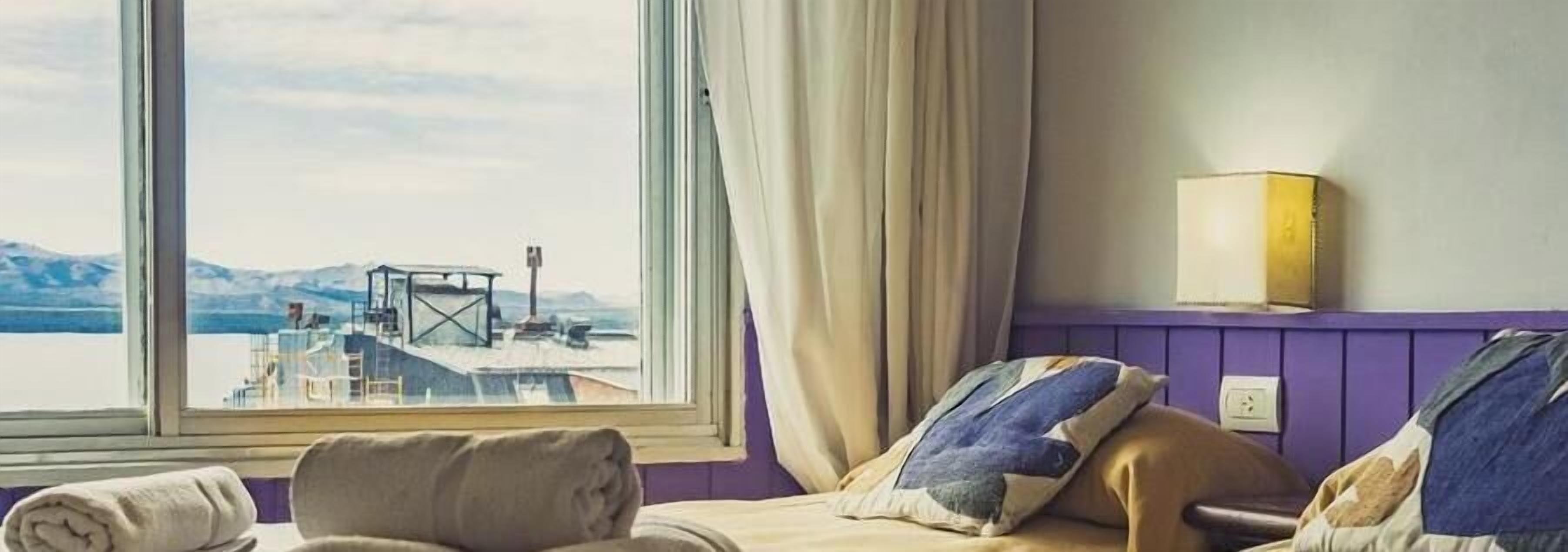 A hotel room with a bed, a window with a view of a dock and mountains, a wall-mounted light, and a purple wall.