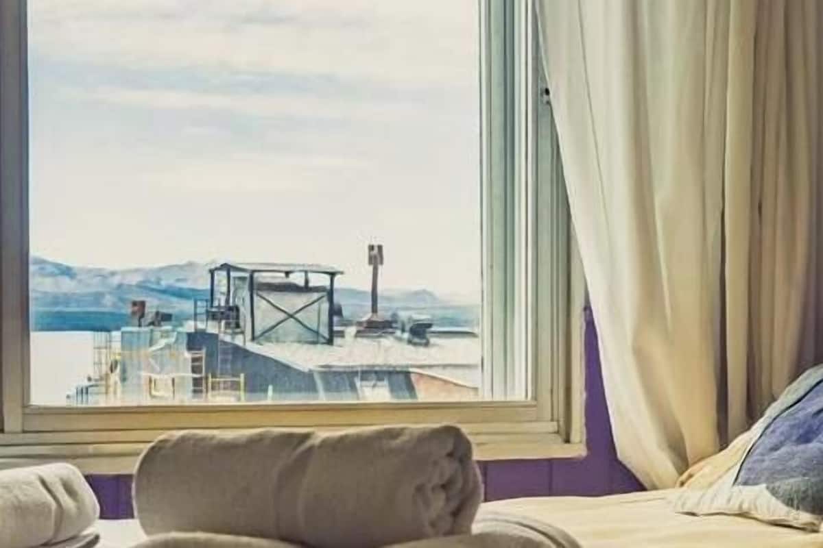Habitación de hotel con cama, ventana que ofrece vistas a un muelle y montañas, una lámpara fijada a la pared y una pared morada.