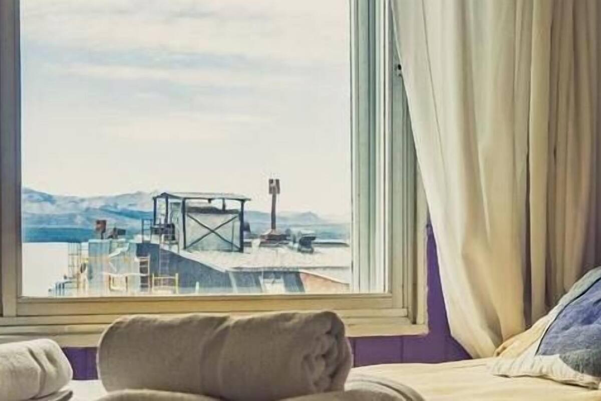 A hotel room with a bed, a window with a view of a dock and mountains, a wall-mounted light, and a purple wall.