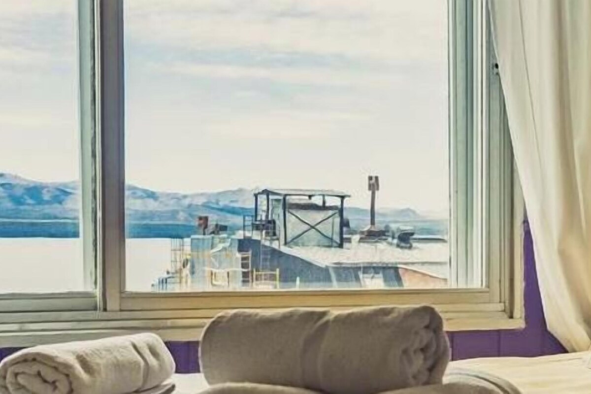 Une chambre d’hôtel avec un lit, une fenêtre donnant sur un quai et des montagnes, une lampe fixée au mur et un mur de couleur violette.