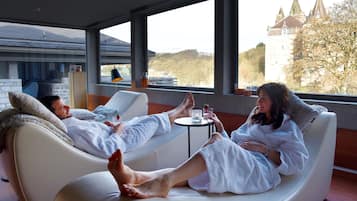 A room with two reclining chairs, a small table, and a view of a castle through large windows.