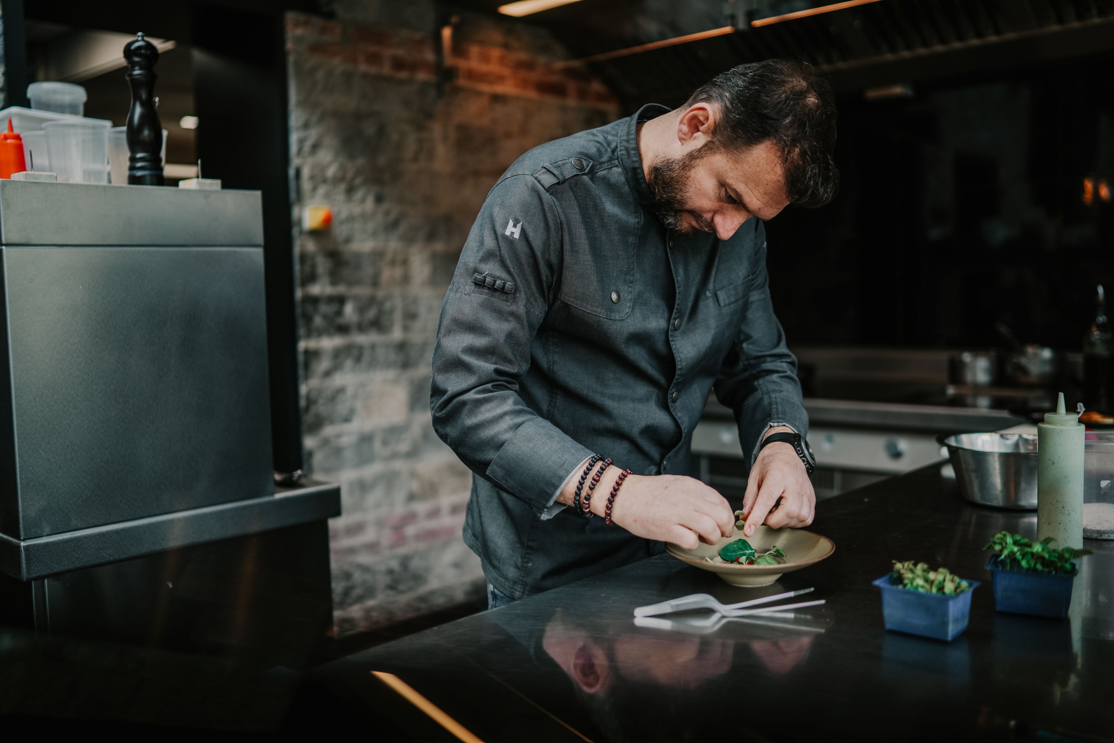 Un chef prepara alimentos en una cocina moderna con pared de ladrillo y electrodomésticos de acero inoxidable.