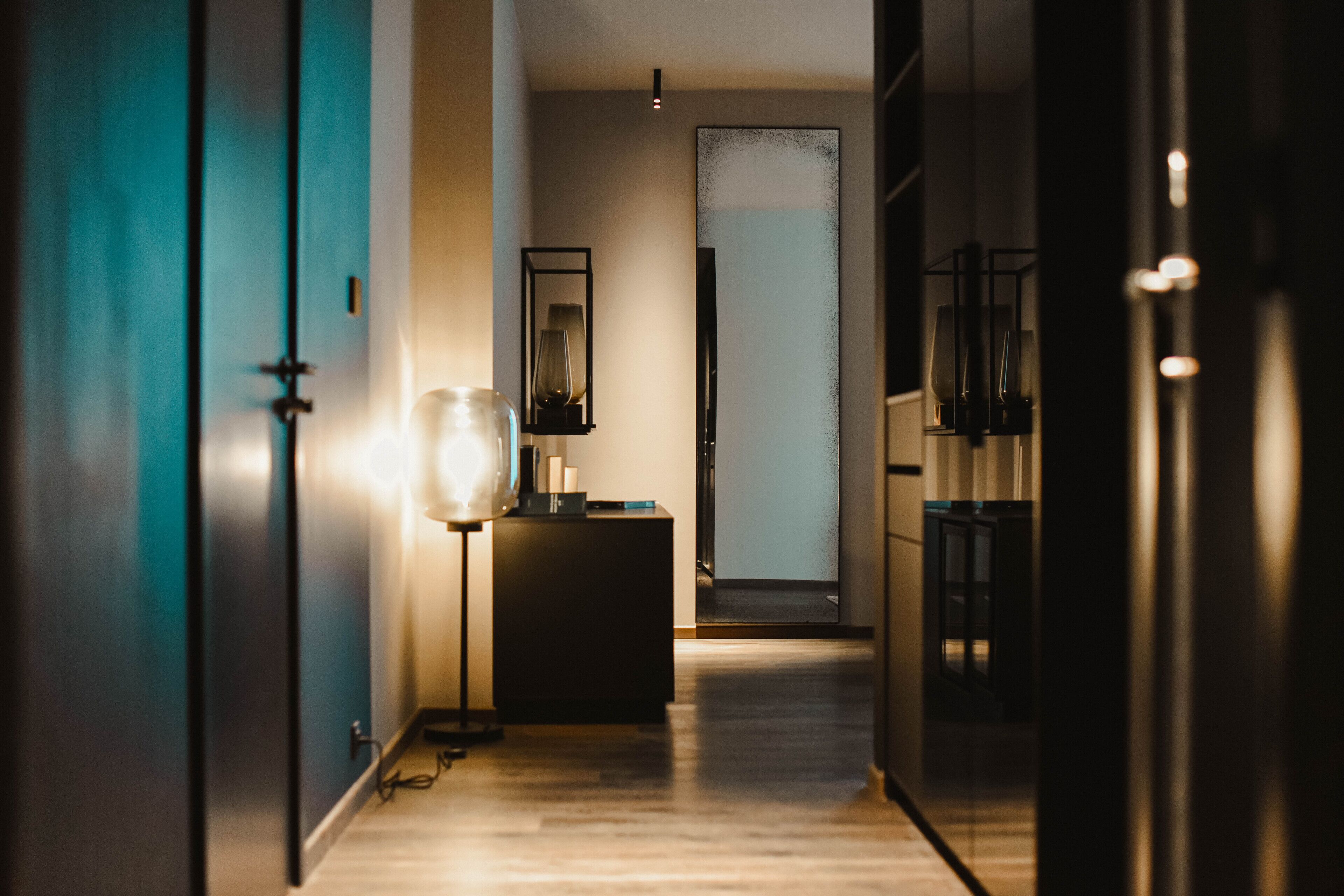 Un couloir moderne avec un plancher en bois, une porte bleue, une console noire surmontée d’une lampe et un grand miroir.