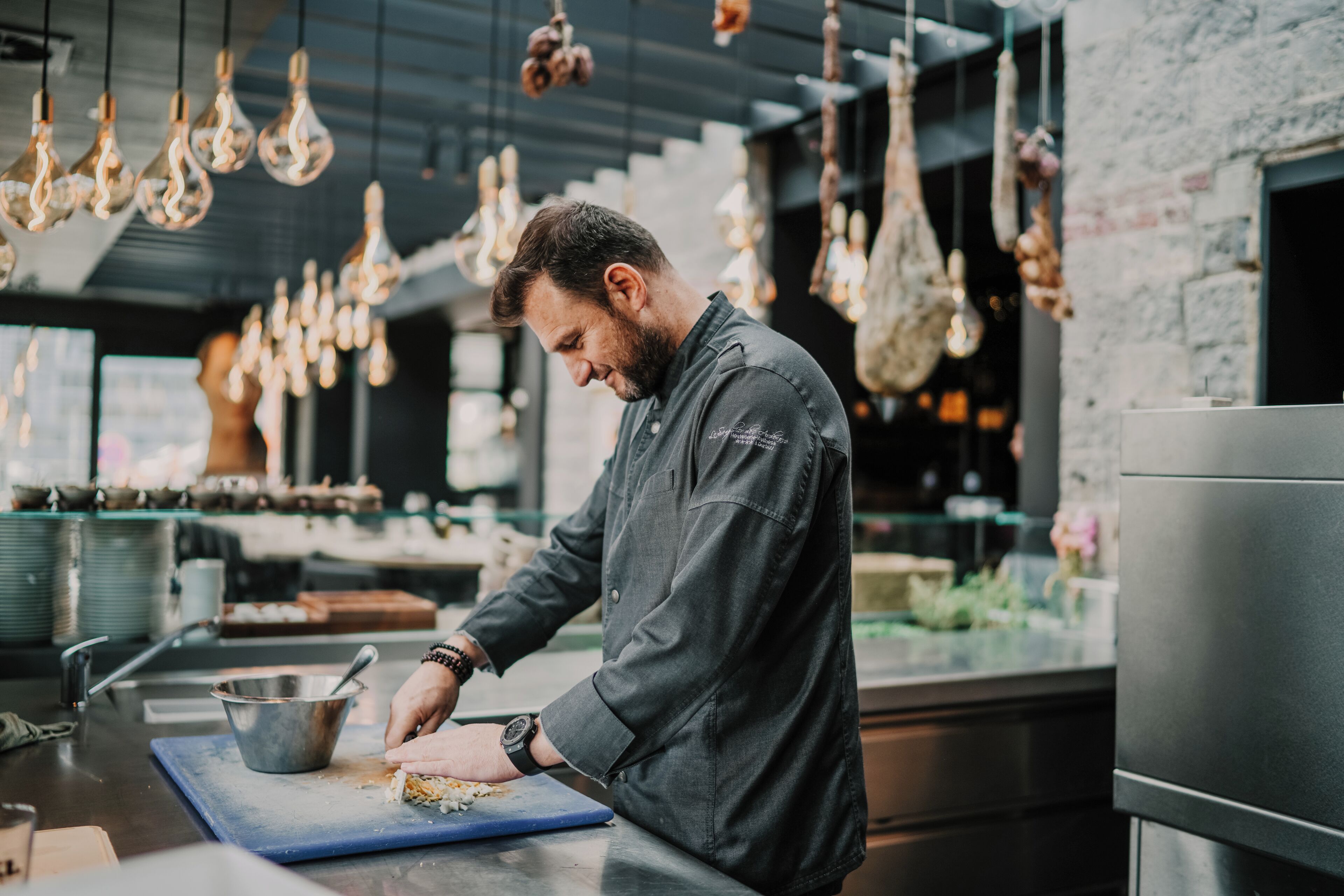 A chef preparing food in a modern kitchen with hanging light fixtures and a stone wall.