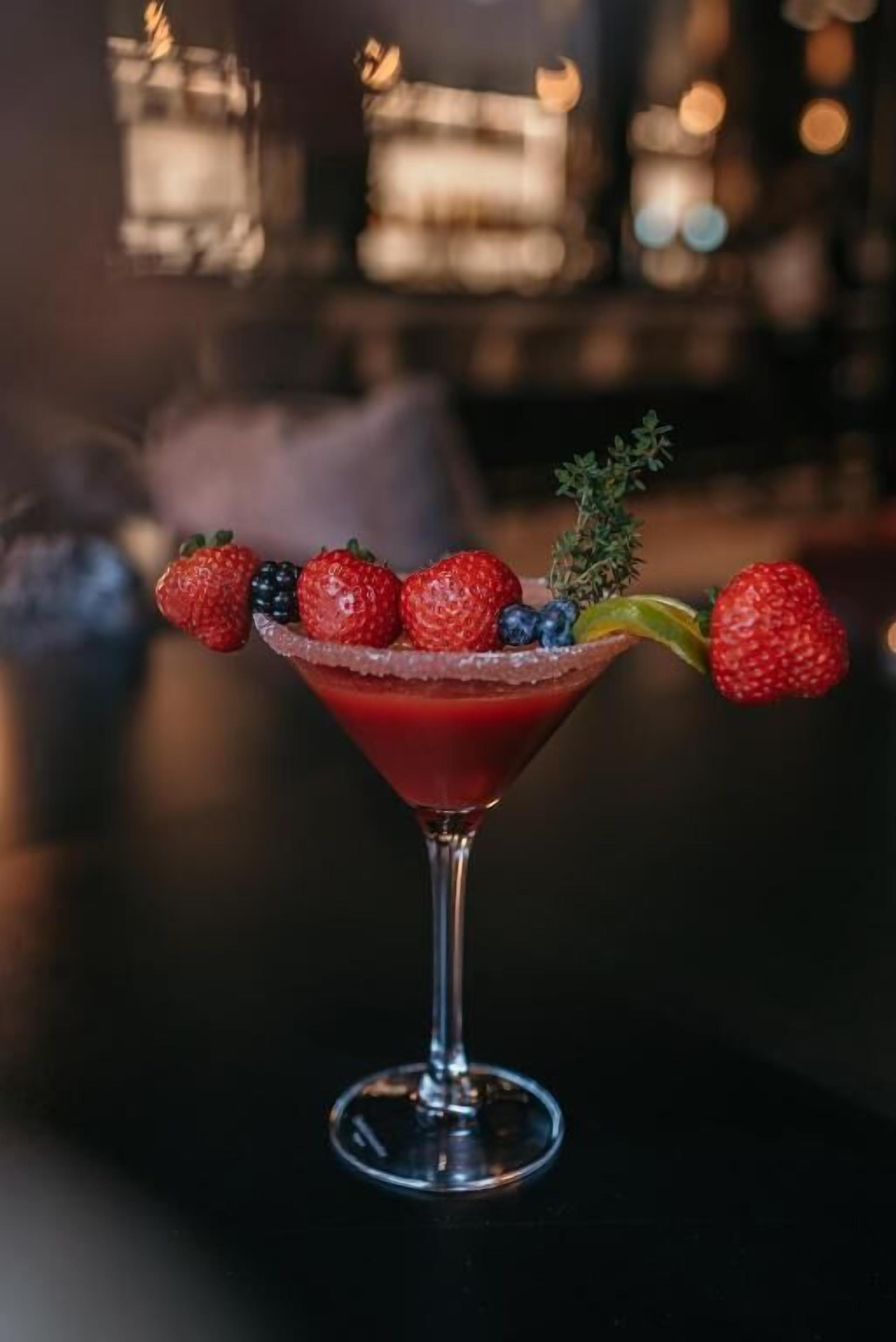 A martini glass with a red drink, garnished with strawberries and blueberries, and a sprig of thyme.