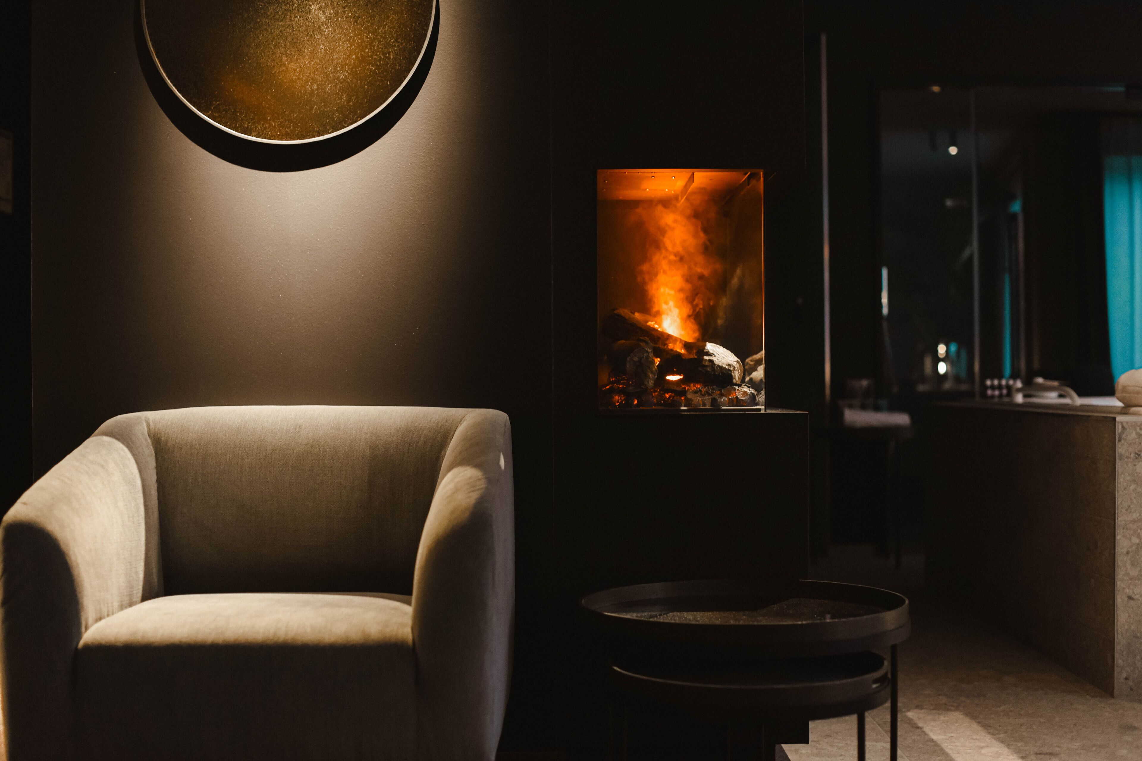 Una sala de estar moderna con un sillón cómodo, una lámpara redonda fijada a la pared y una chimenea con leña y llamas a la vista.