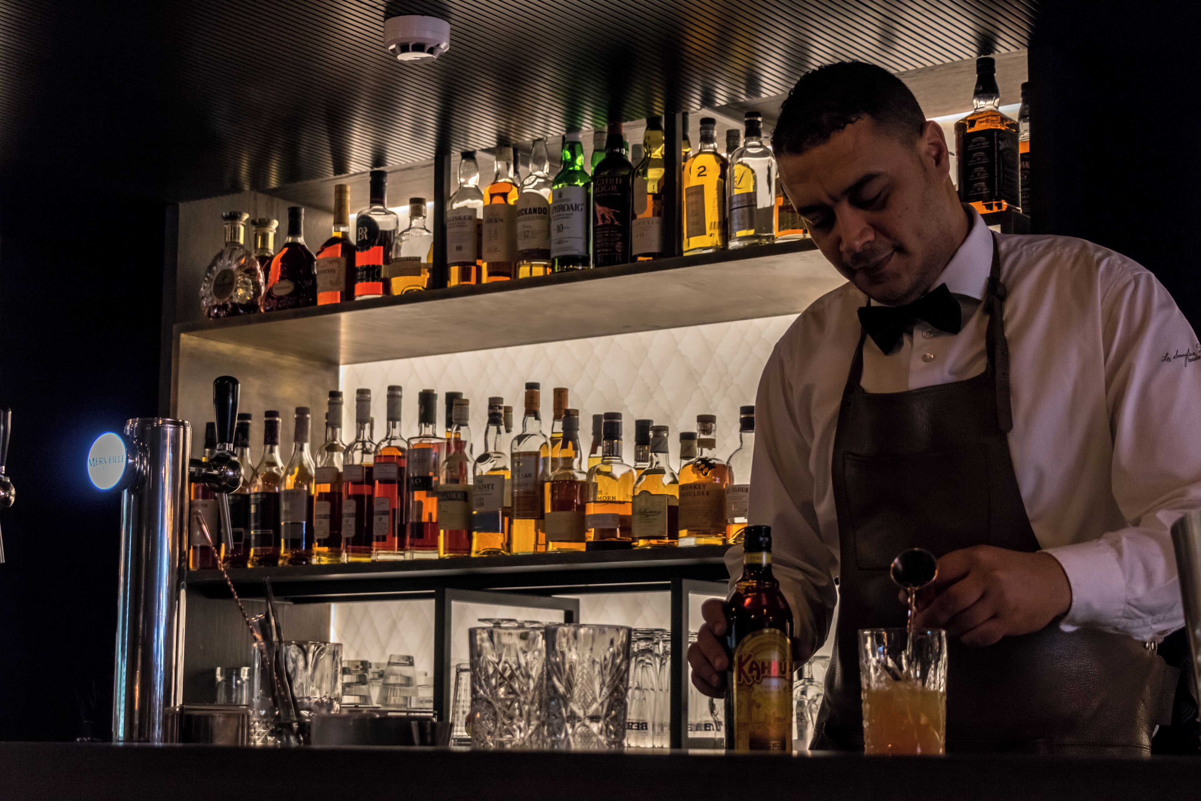 Een bartender bereidt een drankje toe achter een bar met planken vol verschillende drankflessen.