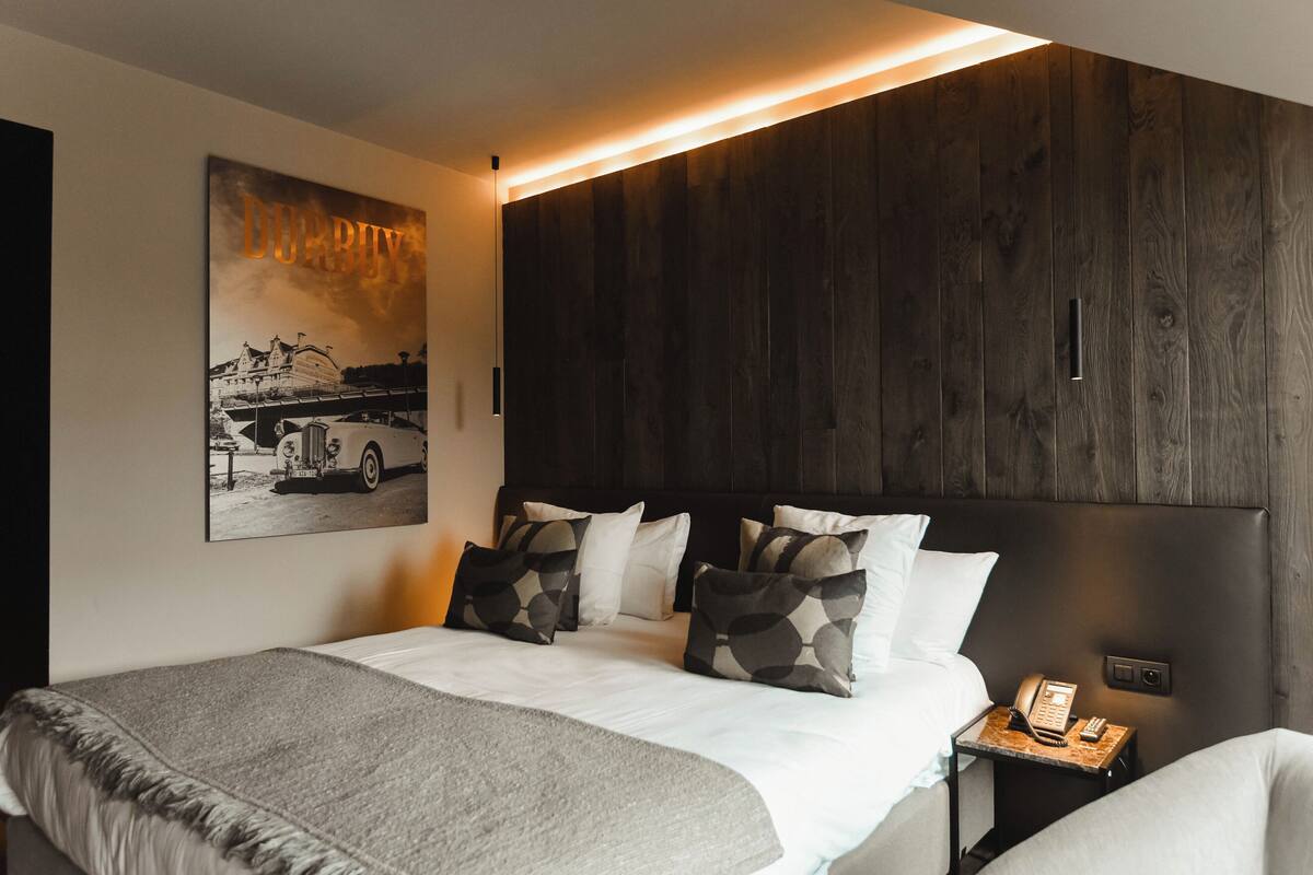 A modern hotel room with a large bed, a headboard with a wooden panel design, a bedside table with a phone, and a wall-mounted artwork depicting a building.